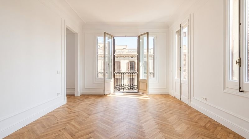 Salón vacío y renovado con paredes blancas, molduras y suelo de madera en espiga, listo para alquilar o vender